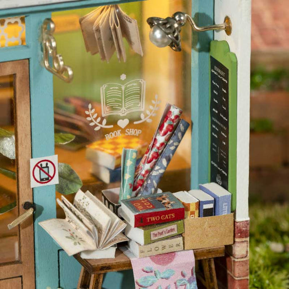 The Free Time Bookshop Model Kit by Robotime showcases a charming miniature bookshop with an inviting doorway. Exterior details include a 'BOOK SHOP' sign with an illustrated book logo, a small lamp, and a no smoking sign. A wooden stand displays an open book alongside various colorful books arranged neatly. Enjoy assembling this DIY kit that captures the essence of a cozy bookshop.