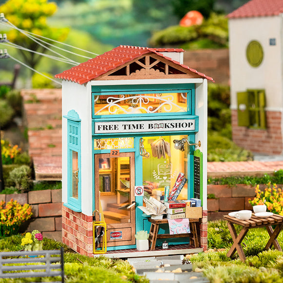 A detailed model kit of the 'Free Time Bookshop' by Robotime, showcasing a colorful facade with blue accents, a red roof, and displayed books. The windows feature miniature items like books, plants, and a chalkboard sign. The scene is set in a charming, grassy landscape with decorative elements around the bookshop, emphasizing the intricate craftsmanship of this DIY kit.