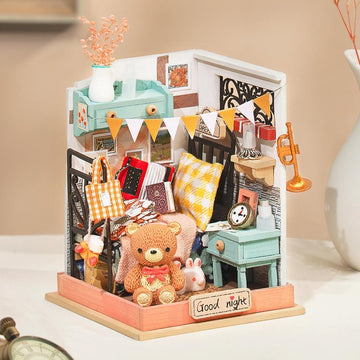The Sweet Dreams Bedroom Model Kit by Robotime features a charming miniature bedroom scene complete with a cozy bed adorned with pillows, a teddy bear, a clock, books, and decorative items. The walls are decorated with pictures and a colorful banner, creating a delightful and inviting atmosphere perfect for a DIY project.