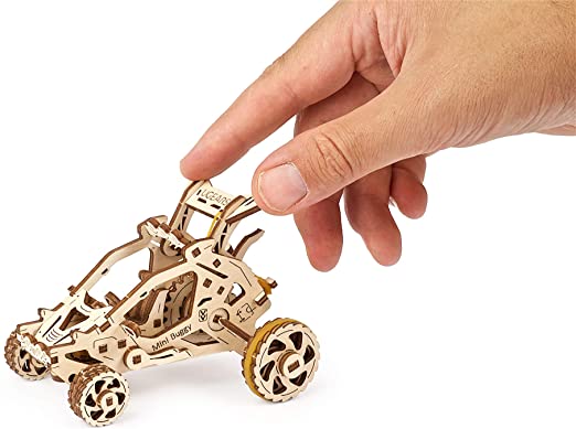 A hand holding a UGears 3D Mini Buggy DIY kit, showcasing the intricate laser-cut wooden design. The buggy features detailed components, including wheels and a driver seat, illustrating the craftsmanship involved in assembling the model.