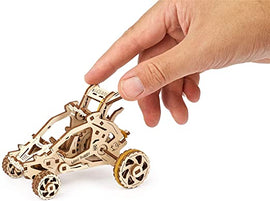A hand holding a UGears 3D Mini Buggy DIY kit, showcasing the intricate laser-cut wooden design. The buggy features detailed components, including wheels and a driver seat, illustrating the craftsmanship involved in assembling the model.