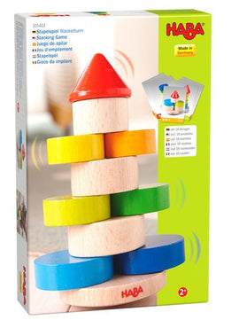 The Stacking Game Wobbly Tower by Haba features colorful wooden pieces in bright shades of red, yellow, green, and blue. The game encourages hand-eye coordination and motor skills as players stack the pieces to form a tower. The packaging shows the product alongside illustrations demonstrating gameplay, suitable for toddlers aged 2 and up. Made in Germany.