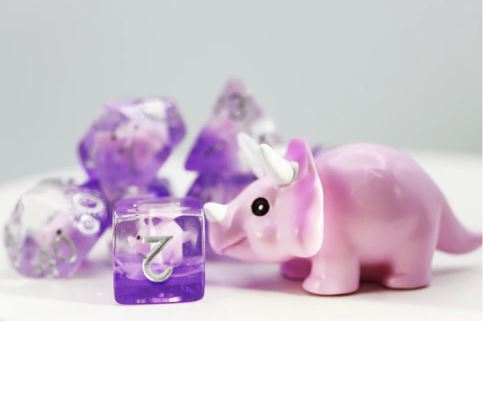 A vibrant Pink Triceratops RPG Dice Set featuring purple, sparkly dice with a small pink triceratops figurine in the foreground. The dice are polyhedral and each has numbers clearly visible, ideal for tabletop role-playing games.