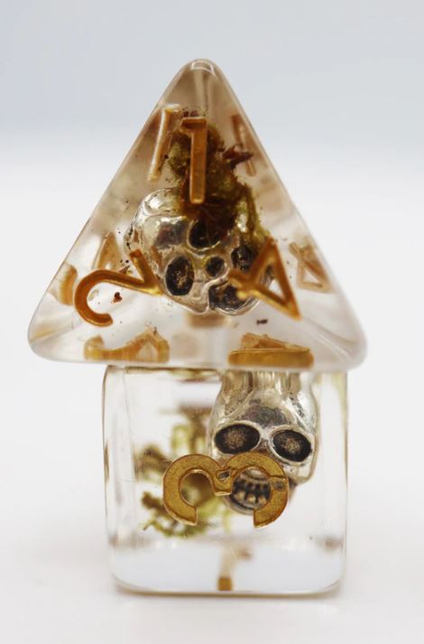 A striking Silver Pirate Skull RPG Dice Set from Foam Brain Games, featuring intricately designed dice with a pirate skull motif. The set includes various dice shapes encased in a transparent resin, adorned with gold accents. Perfect for tabletop RPG enthusiasts looking to enhance their gaming experience with a unique and thematic accessory.