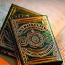 A close-up view of the High Victorian Card Deck by Theory 11 displays its intricate gold and green design. The playing cards are elegantly designed with detailed motifs and a luxurious finish, positioned atop an open book that adds a scholarly touch.