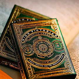 A close-up view of the High Victorian Card Deck by Theory 11 displays its intricate gold and green design. The playing cards are elegantly designed with detailed motifs and a luxurious finish, positioned atop an open book that adds a scholarly touch.