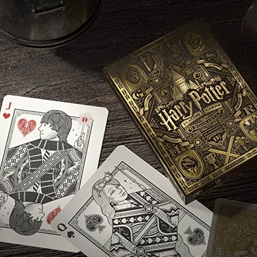 A close-up of a Harry Potter-themed playing card deck from Theory 11. The deck features intricate gold and black illustrated designs with iconic symbols, including a jack and queen of hearts. The scene is set on a textured wooden surface, enhancing the magical atmosphere.
