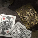 A close-up of a Harry Potter-themed playing card deck from Theory 11. The deck features intricate gold and black illustrated designs with iconic symbols, including a jack and queen of hearts. The scene is set on a textured wooden surface, enhancing the magical atmosphere.