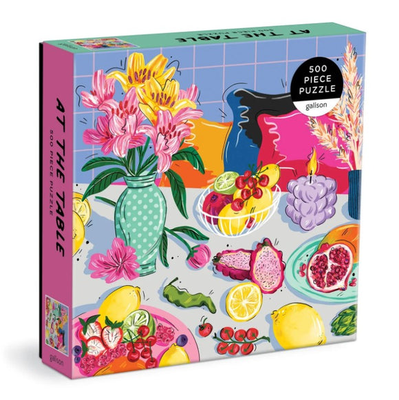 Colorful jigsaw puzzle titled 'At the Table' by Galison featuring a vibrant table setting with flowers, fruits, and decorative items. The puzzle contains 500 pieces, ideal for family fun and relaxation.