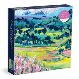 Galison Painted Hills 500 Piece Jigsaw Puzzle featuring a vibrant landscape with rolling hills, meadows, and colorful trees. The box showcases a bright and artistic design, perfect for puzzle enthusiasts and art lovers.