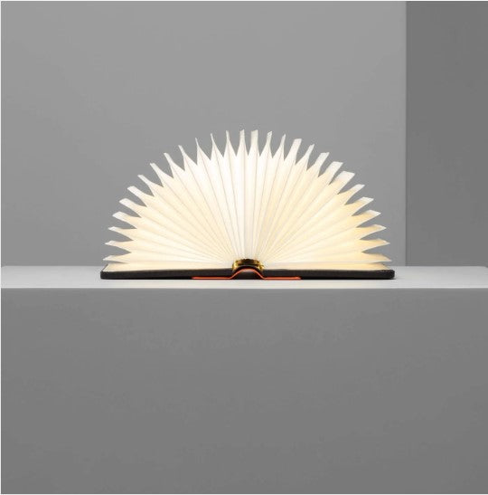 Lito Mini Orange/Black Charging Book by Lumio displayed on a shelf. The book is open, resembling a fan shape with illuminated pages, perfect for reading or as a stylish decorative light.
