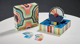 Wavelength game by Palm Court displayed with its vibrant, multi-colored packaging and game components, including a circular dial and colorful cards laid out on a table. The design features bold, psychedelic patterns that enhance its visual appeal, inviting players to engage in fun and strategic gameplay.