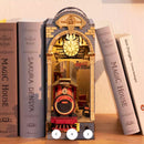 The Time Travel DIY Book Nook by Robotime showcases a beautifully crafted miniature scene featuring a vintage red train inside an archway structure. The design includes intricate details like a clock, welcoming sign, and a cozy interior, perfectly placed among books on a shelf.