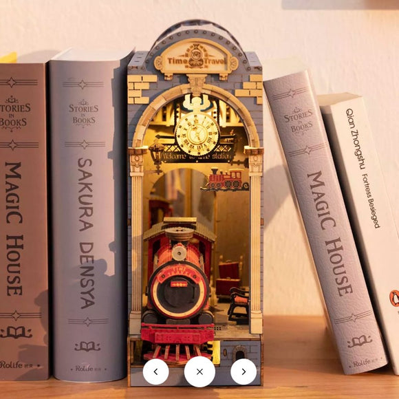 The Time Travel DIY Book Nook by Robotime showcases a beautifully crafted miniature scene featuring a vintage red train inside an archway structure. The design includes intricate details like a clock, welcoming sign, and a cozy interior, perfectly placed among books on a shelf.