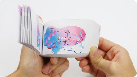 A person holding the Simbiosis Carnal flipbook by FlipBoku, showcasing colorful illustrations of abstract figures in playful poses. The flipbook captures dynamic movement with each page, highlighting its artistic design.