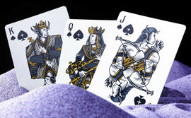 A close-up image of three playing cards from the Jupiter Planets set by VPC, featuring the King, Queen, and Jack of Spades. The cards showcase intricate designs with striking colors, set against a backdrop of purple sand, emphasizing their artistic details.