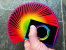 A vibrant deck of Unicorn Playing Cards, featuring a rainbow gradient design radiating from the center, with a smooth black background. A hand holds a small section of the fanned-out cards, showcasing their colorful edges.