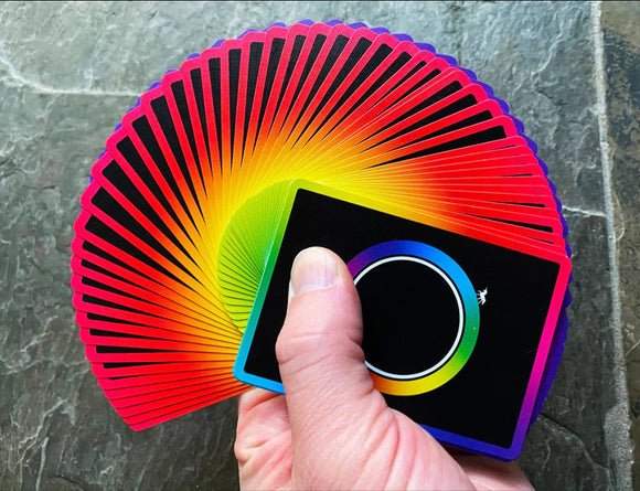 A vibrant deck of Unicorn Playing Cards, featuring a rainbow gradient design radiating from the center, with a smooth black background. A hand holds a small section of the fanned-out cards, showcasing their colorful edges.