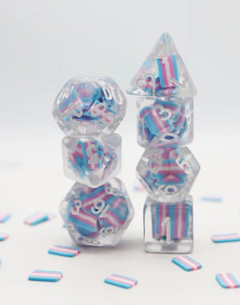 A visually striking Transgender Flag RPG Dice Set by Foam Brain, featuring transparent dice in various shapes—such as polyhedrons—each showcasing vibrant pink, blue, and white colors reminiscent of the transgender pride flag. The dice are arranged in a stacked formation with smaller dice at the bottom and larger ones at the top, creating a colorful display against a soft, light background. Scatterings of colored pieces, reflecting the flag's colors, are positioned around the base.