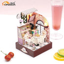 A beautifully crafted DIY miniature Sweet Cake Station featuring a pastel-colored pastry shop with a display case of cakes, two chairs, a table with a lace cloth, and decorative elements like flowers and a window. The scene is charming and inviting, perfect for those who enjoy crafting and creating detailed models.