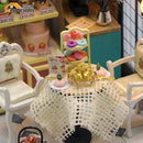 A detailed miniature cake station DIY kit featuring a beautifully arranged table with a lace tablecloth, two decorative chairs, colorful dessert items including mini cakes, macarons, and a display stand, all set in a charming café-like scene. Perfect for crafting enthusiasts and collectors.