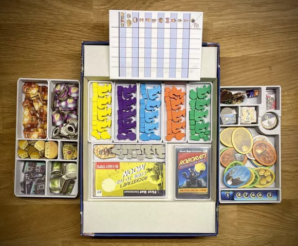 Open box of the game 'First Rat' by Pegasus Spiele showing organized game components including colorful tokens, cards, and game board. The box features sections filled with various game pieces such as dice, cards, and coins, as well as a player score sheet.