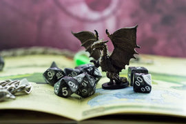 A close-up view of Eureka! D&D Adult One Shots game pieces, featuring a dragon miniature poised among various gaming dice and an open adventure book. The scene showcases a vibrant tabletop gaming setup, perfect for Dungeons & Dragons enthusiasts.