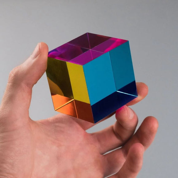 A colorful, reflective cube held in a person's hand, designed by CMY Cubes. The cube features vibrant colors of blue, purple, yellow, and orange, showcasing a modern and artistic design, ideal for puzzle enthusiasts and collectors.