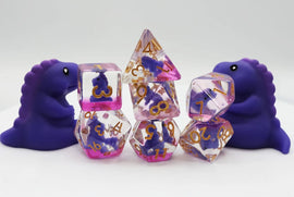 A vibrant Purple T-Rex RPG Dice Set by Foam Brain featuring various dice in purple and pink hues with gold numbering. Two cute purple T-Rex figures are positioned beside the dice, enhancing the playful and whimsical appeal of the game set.