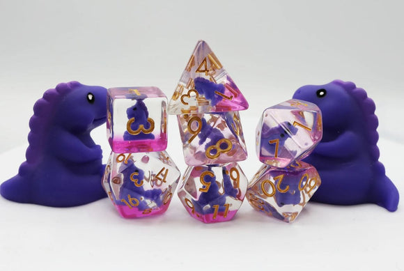 A vibrant Purple T-Rex RPG Dice Set by Foam Brain featuring various dice in purple and pink hues with gold numbering. Two cute purple T-Rex figures are positioned beside the dice, enhancing the playful and whimsical appeal of the game set.
