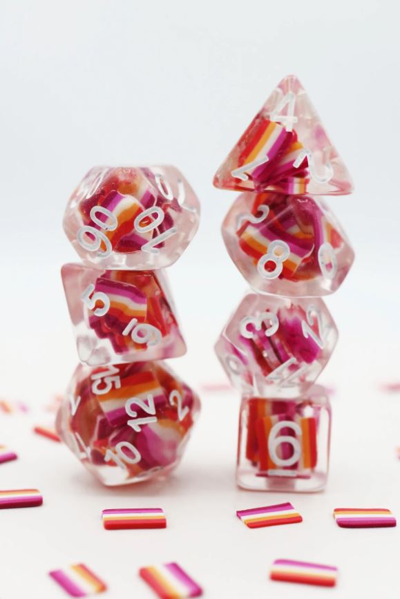 A colorful set of Community Lesbian Flag RPG dice by Foam Brain. The dice feature vibrant pink, orange, and white stripes encased in crystal-clear resin, with clear white numbers printed on them. The arrangement displays a variety of shapes including a d20, d12, and d6, set against a background of small rainbow strips.