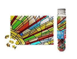 Colorful stained glass window design jigsaw puzzle with vibrant pieces scattered around. The puzzle showcases intricate patterns with reds, greens, and yellows, reflecting a sunlight effect. A clear container filled with additional jigsaw pieces is shown beside the completed segment.