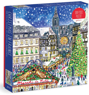 Christmas in France jigsaw puzzle by Michael Storrings, featuring a vibrant and festive winter scene with a Christmas market and snow-covered streets. The puzzle contains 500 pieces and showcases charming architecture, a large decorated Christmas tree, and bustling holiday shoppers.