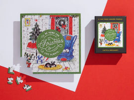 A beautifully illustrated jigsaw puzzle titled 'I'll Be Home for Christmas, Like Always,' featuring a cozy holiday scene with a decorated Christmas tree, a blue chair, and sleeping cats on a rug. The 100-piece puzzle is shown partially assembled on a white background, with its compact black box displayed nearby.
