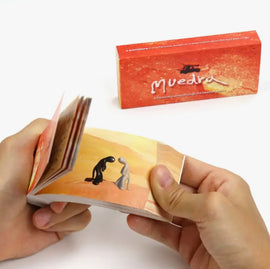 A person holding a colorful flipbook titled 'Muedra' by Boku Flipbooks. The flipbook features animated illustrations of two characters in a desert landscape, showcasing a unique storytelling experience through its pages.