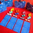 A colorful game setup for Break the Cube by Iello, featuring various geometric puzzle pieces in vibrant colors arranged alongside strategy cards on a red background. The game includes several uniquely shaped blocks, game boards with designs, and cards with numbers and symbols, encouraging creative problem-solving and teamwork.