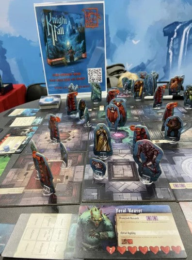 A close-up view of the tabletop game 'Knight Fall' by RED RAVEN GAMES. The game board features various colorful character figurines and player cards, creating an immersive fantasy setting. The backdrop displays the game's vibrant artwork and promotional details.