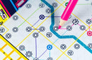 Close-up view of the game 'Next Station London' featuring a colorful game board with geometric symbols and a pink pencil marking the paths. The vibrant design captures the excitement of strategic gameplay as players navigate London's transit system.