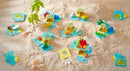 The Flotsam Float game by Haba is showcased on a sandy surface. It features colorful, playful components such as wooden pieces shaped like boats, animals, and other beach-themed items, arranged on light blue game cards. The scene is set with natural elements like seashells and green leaves, evoking a fun, outdoor play environment.