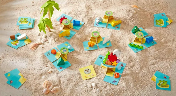 The Flotsam Float game by Haba is showcased on a sandy surface. It features colorful, playful components such as wooden pieces shaped like boats, animals, and other beach-themed items, arranged on light blue game cards. The scene is set with natural elements like seashells and green leaves, evoking a fun, outdoor play environment.