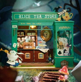 A detailed DIY model kit of Alice's Tea Store featuring a miniature green shop exterior. The shop has a sign that reads 'ALICE TEA STORE' and various decorative elements, including a mirror, teacups, and playing cards. Inside, there are whimsical figures and an array of teacups, adding to the fantasy theme reminiscent of Alice in Wonderland.