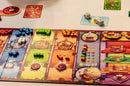A colorful board game setup featuring various colored cubes, dice, and player cards spread across a game board. Players interact with game elements themed around fantasy characters and events.