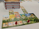 A colorful board game setup featuring The Palaces of Carrara by GAME BREWER LLC. The game includes a large building model and various card pieces on a green and illustrated board, with red player pieces positioned on the board tracks. The game elements showcase vibrant artwork and a strategic layout for gameplay.