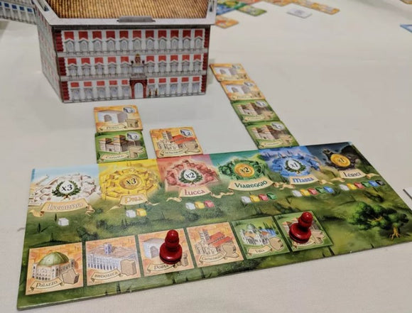 A colorful board game setup featuring The Palaces of Carrara by GAME BREWER LLC. The game includes a large building model and various card pieces on a green and illustrated board, with red player pieces positioned on the board tracks. The game elements showcase vibrant artwork and a strategic layout for gameplay.