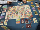 A tabletop view of the KuZOOka board game by Pegasus Spiele. The game board features colorful numbered spaces and player tokens in various colors. Surrounding the board are game cards and resource tokens, with players interacting and strategizing during gameplay.