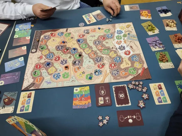 A tabletop view of the KuZOOka board game by Pegasus Spiele. The game board features colorful numbered spaces and player tokens in various colors. Surrounding the board are game cards and resource tokens, with players interacting and strategizing during gameplay.