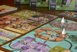 Four Humours game by Adam's Apple Games displayed on a wooden table. The game boards feature colorful paths and various locations marked, surrounded by player pieces and tokens.