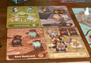 Four Humours game by Adam's Apple Games featuring colorful game boards labeled Sacred Swamp, Laboratory, Bard Boulevard, and Cybal's Cavern. Each board displays quirky characters and game markers, creating an engaging and vibrant gaming experience.