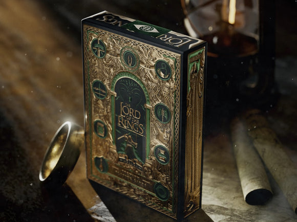 A beautifully illustrated Lord of the Rings card deck by Theory 11. The box features intricate gold detailing, iconic symbols from the series, and a rich green background, set against a softly lit wooden surface with a vintage compass and scrolls.