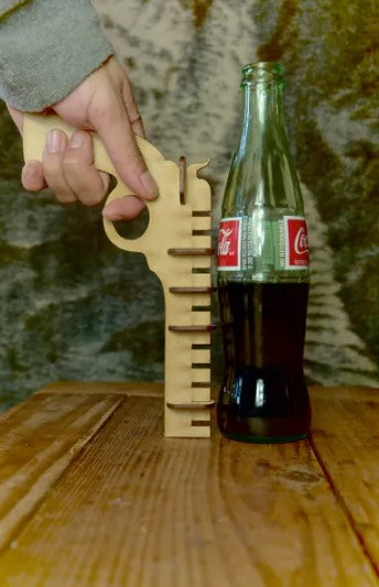 A hand holding a unique wooden bottle opener shaped like a gun, positioned next to a Coca-Cola bottle on a wooden table. The opener features measurement markings along the side, adding a playful design element to this functional tool.